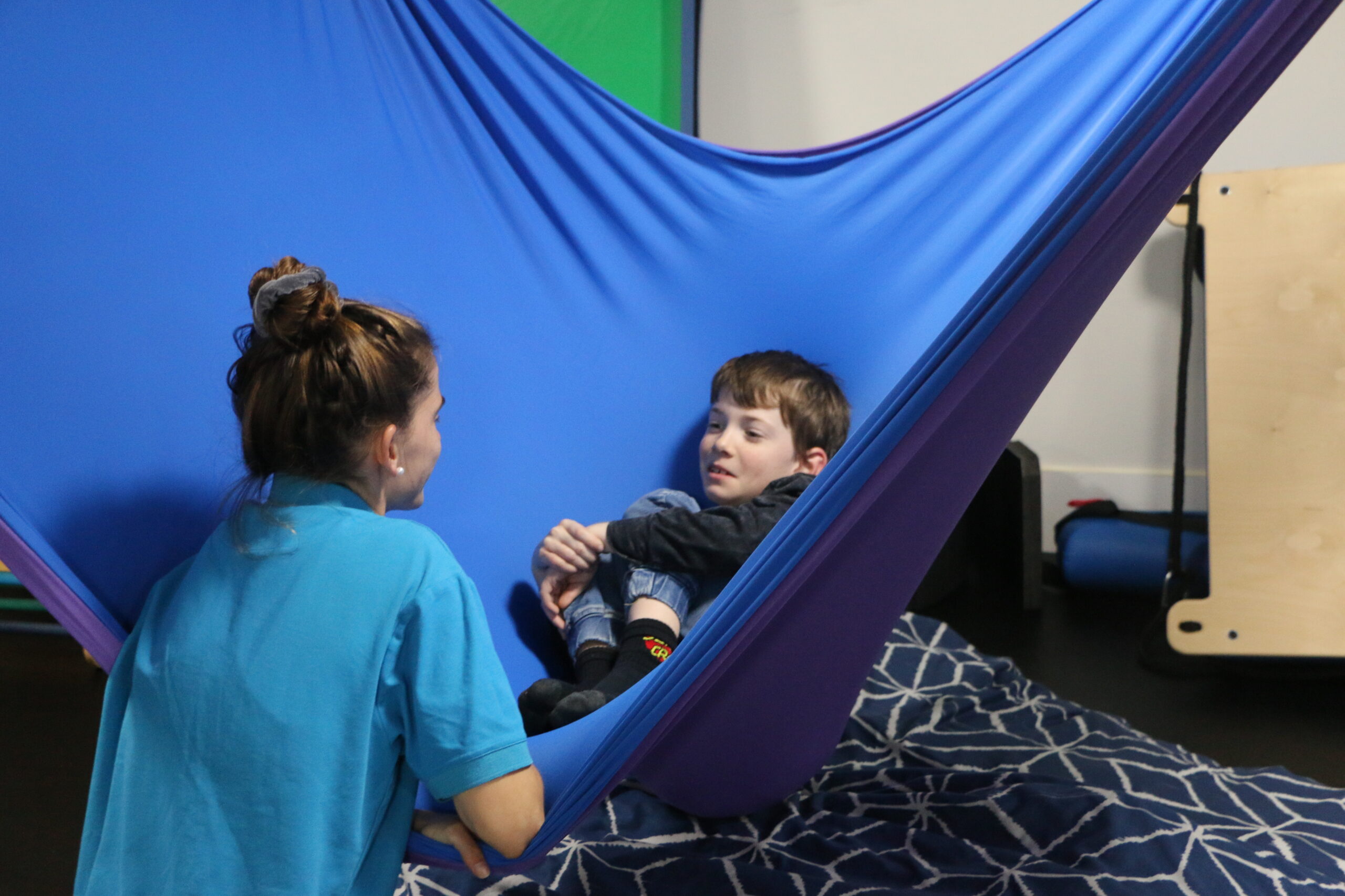 A therapist and child in a blur and purple lycra sensory swing at an occupational therapy clinic.